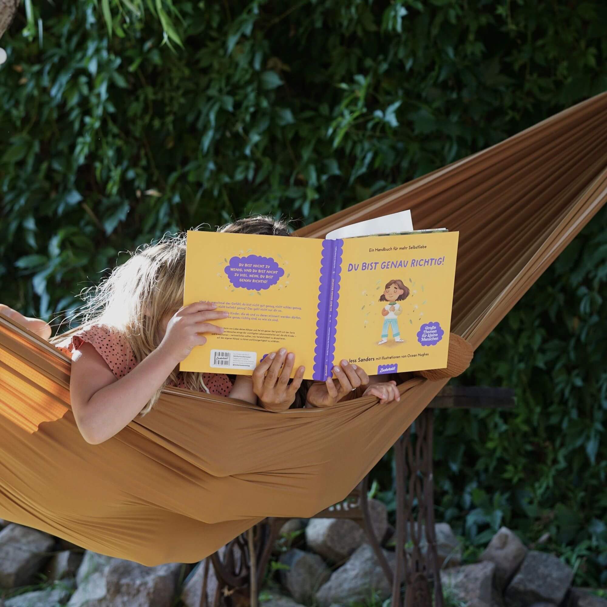 Zwei Kinder entspannen sich gemeinsam in einer braunen Hängematte im Freien und lesen "Ich bin genau richtig" von Jessica Sanders. Umgeben von üppig grünem Laub genießen sie eine friedliche Umgebung, die zu Selbstliebe inspiriert.